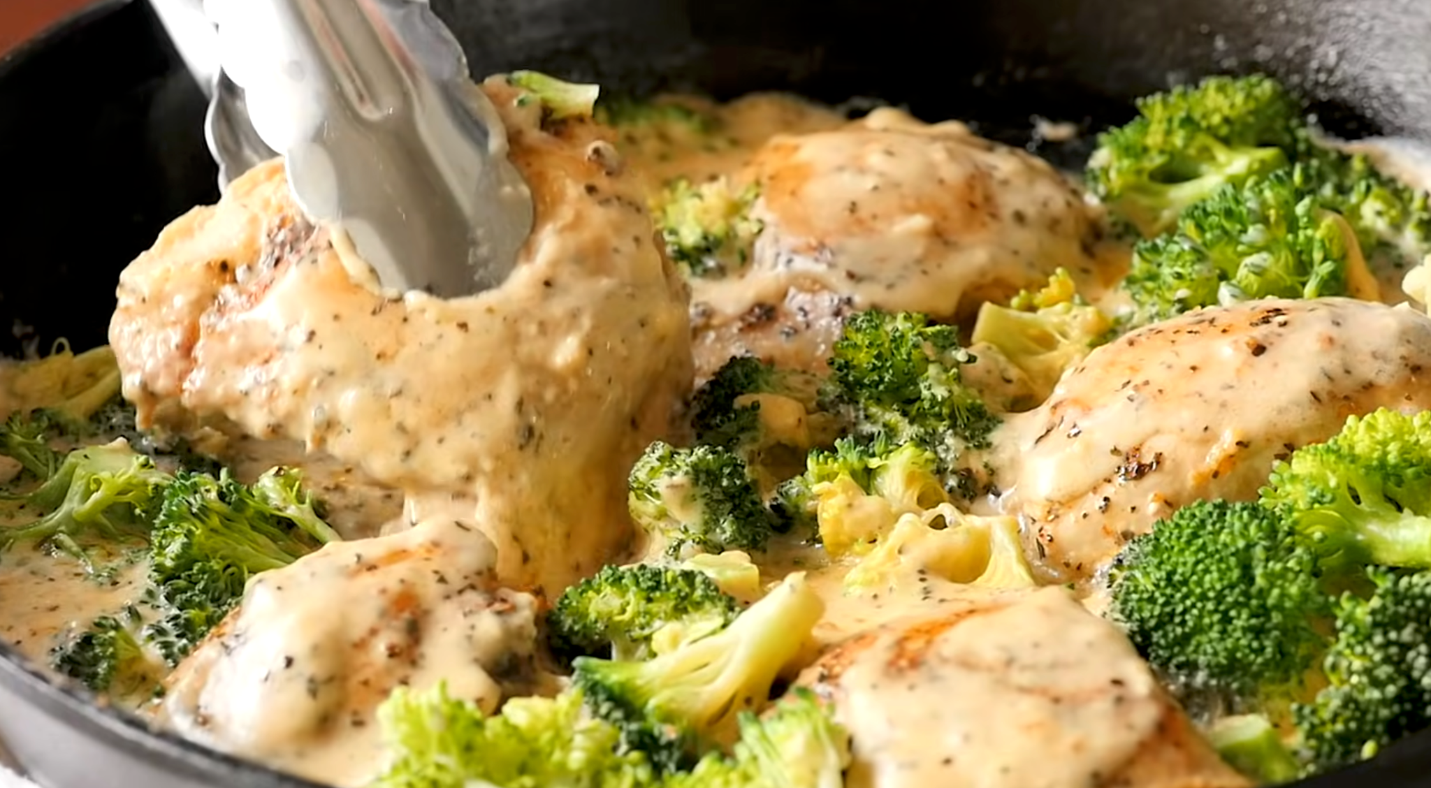 Creamy chicken and broccoli being turned in the skillet with kitchen tongs