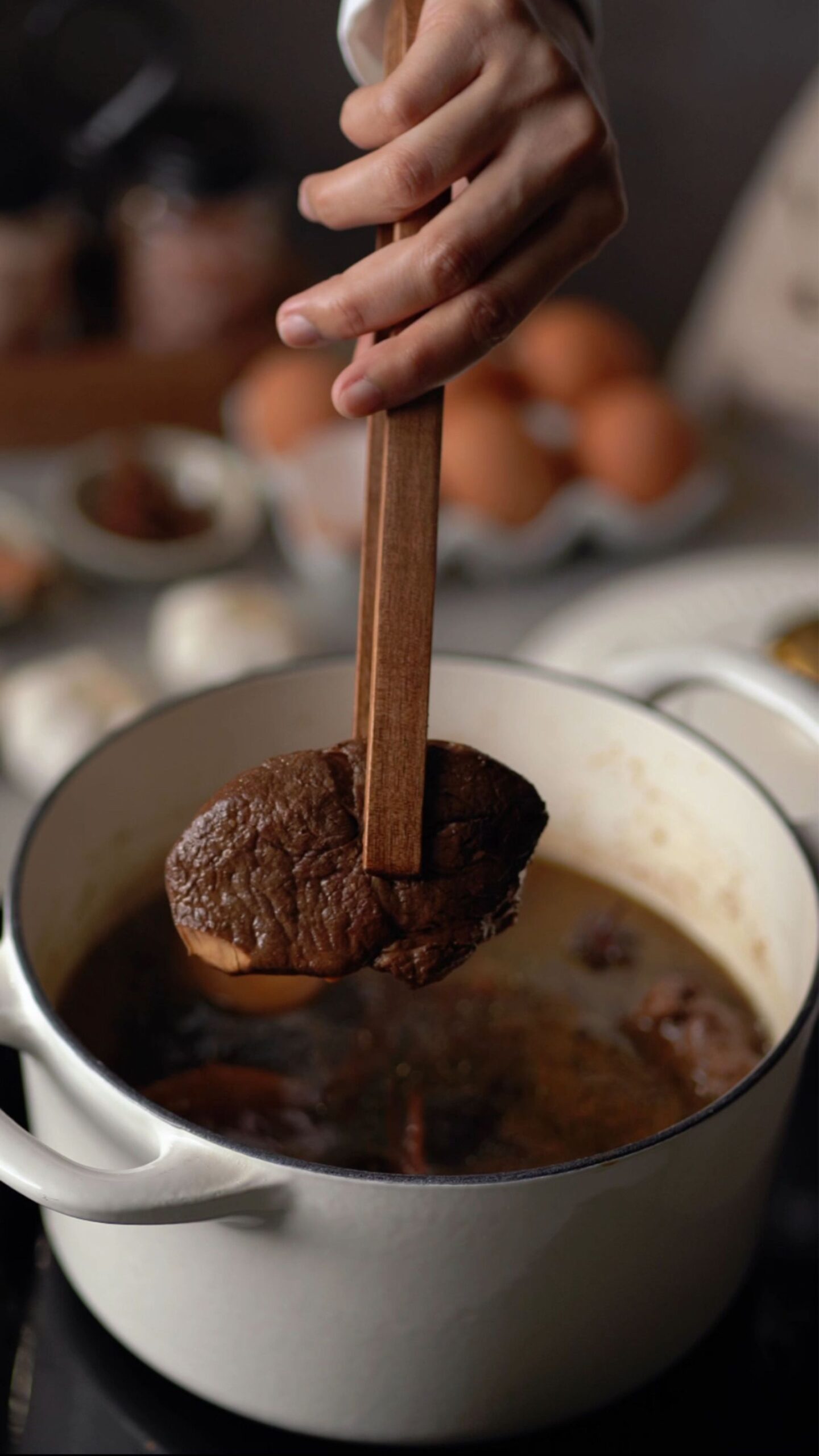 A hand lifting tender braised beef from a pot of birria broth with tongs