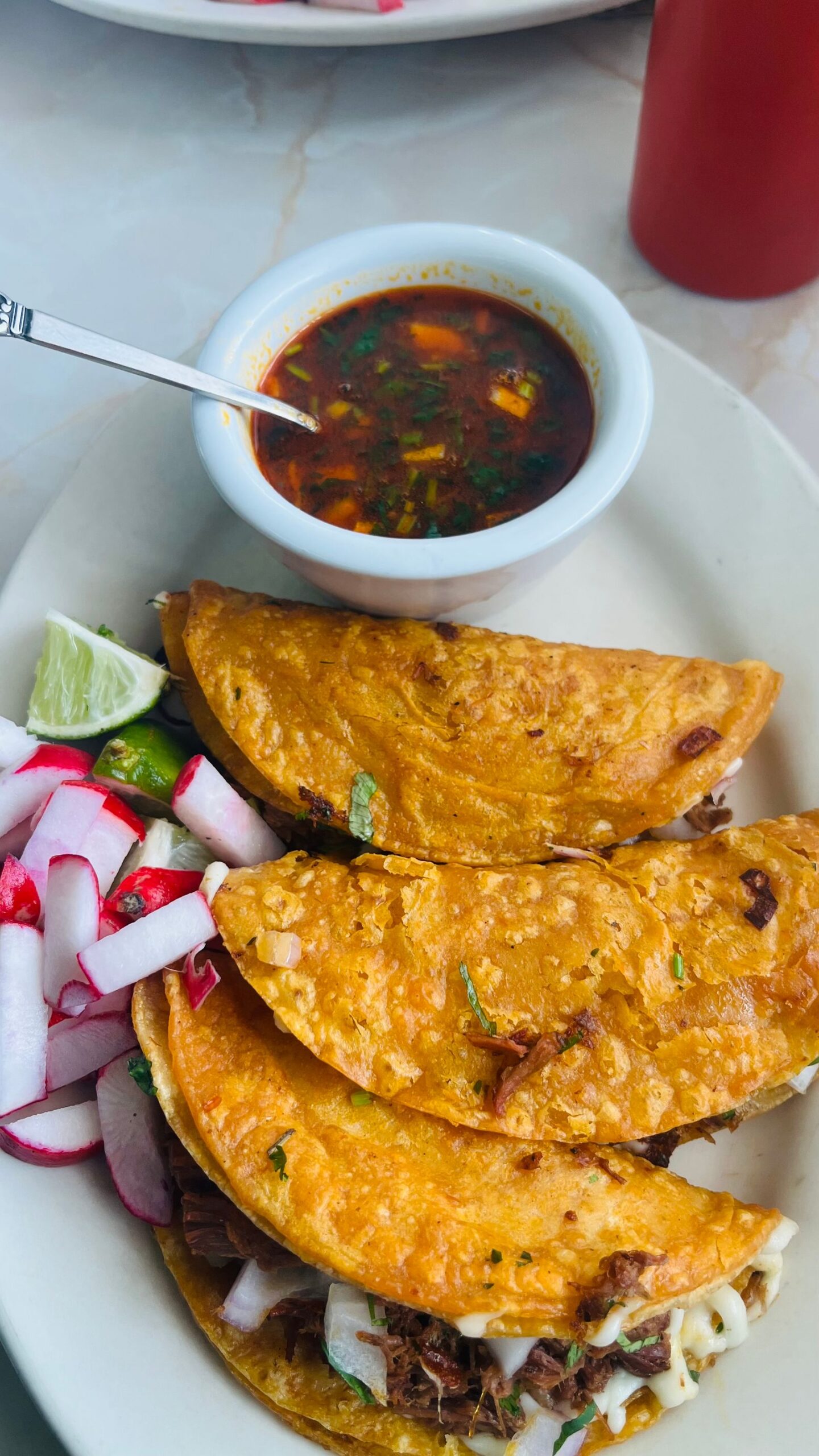 Crispy birria tacos on a plate with a bowl of beef consomm&eacute; for dipping, radishes and lime