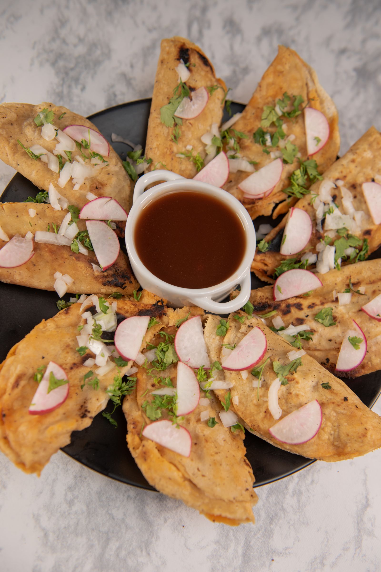 A plate of birria tacos arranged around a bowl of consomm&eacute; for dipping